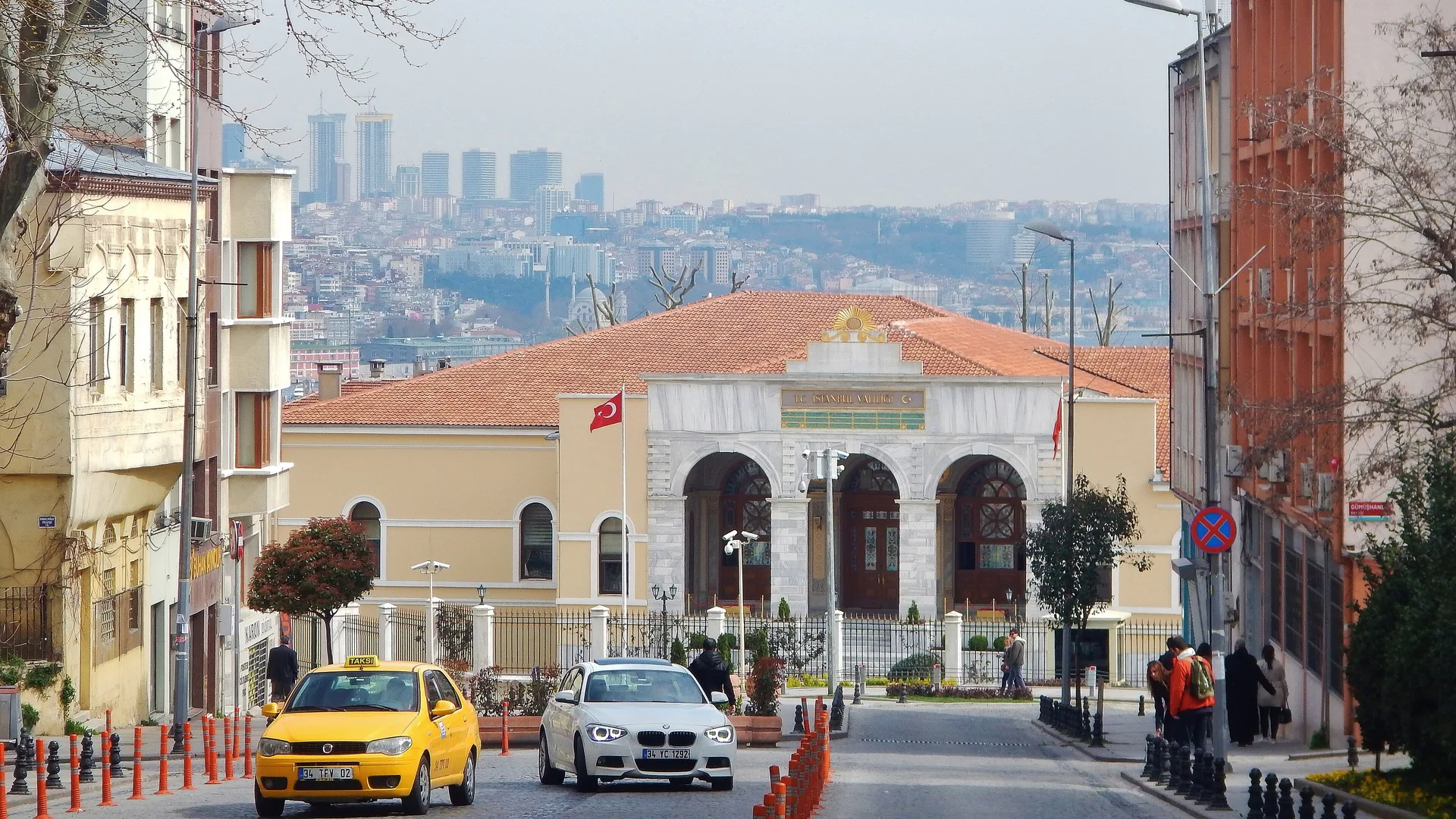 Kırmızı kiremit çatılı, kemerli tarihi bir yapı; önünde taksi ve beyaz otomobil