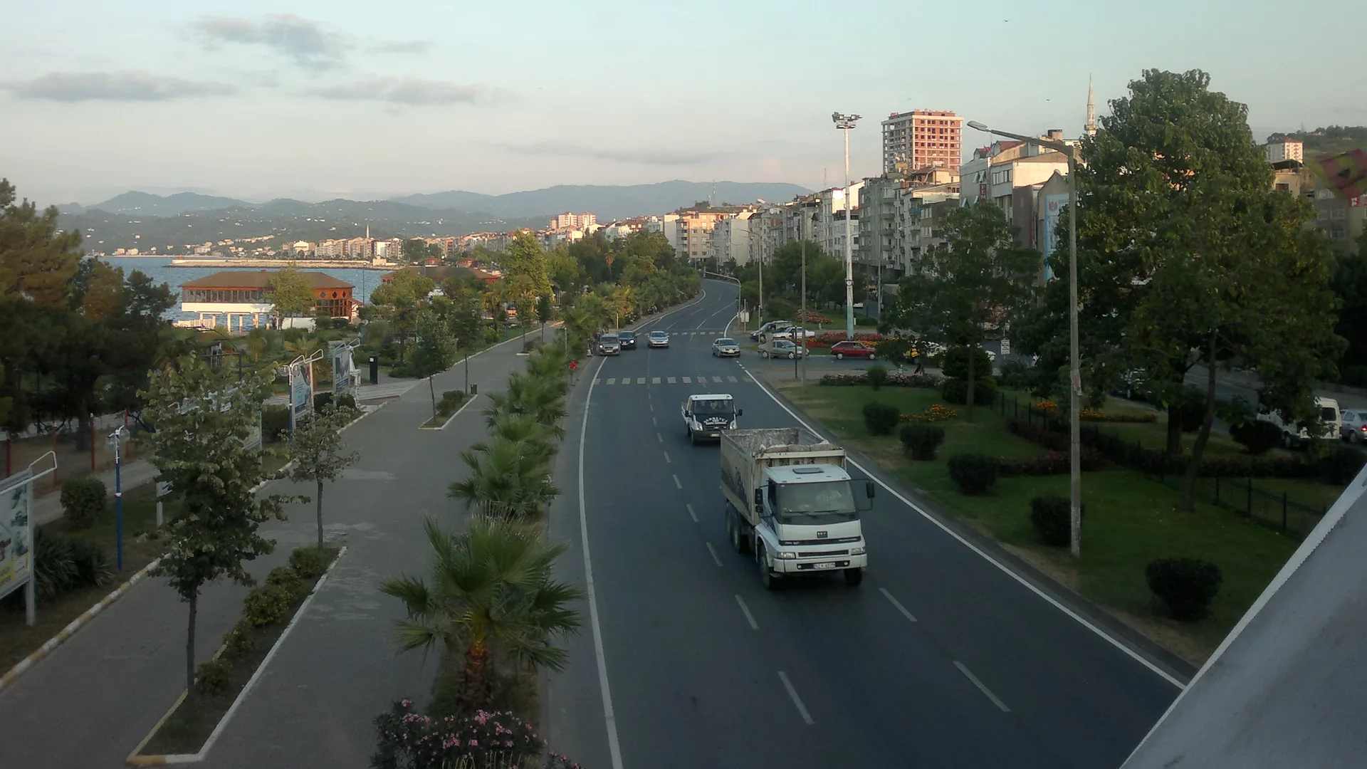 Deniz kıyısında ağaçlı bir sahil yolu, yolda giden kamyon ve araçlar, arkada binalar ve dağlar