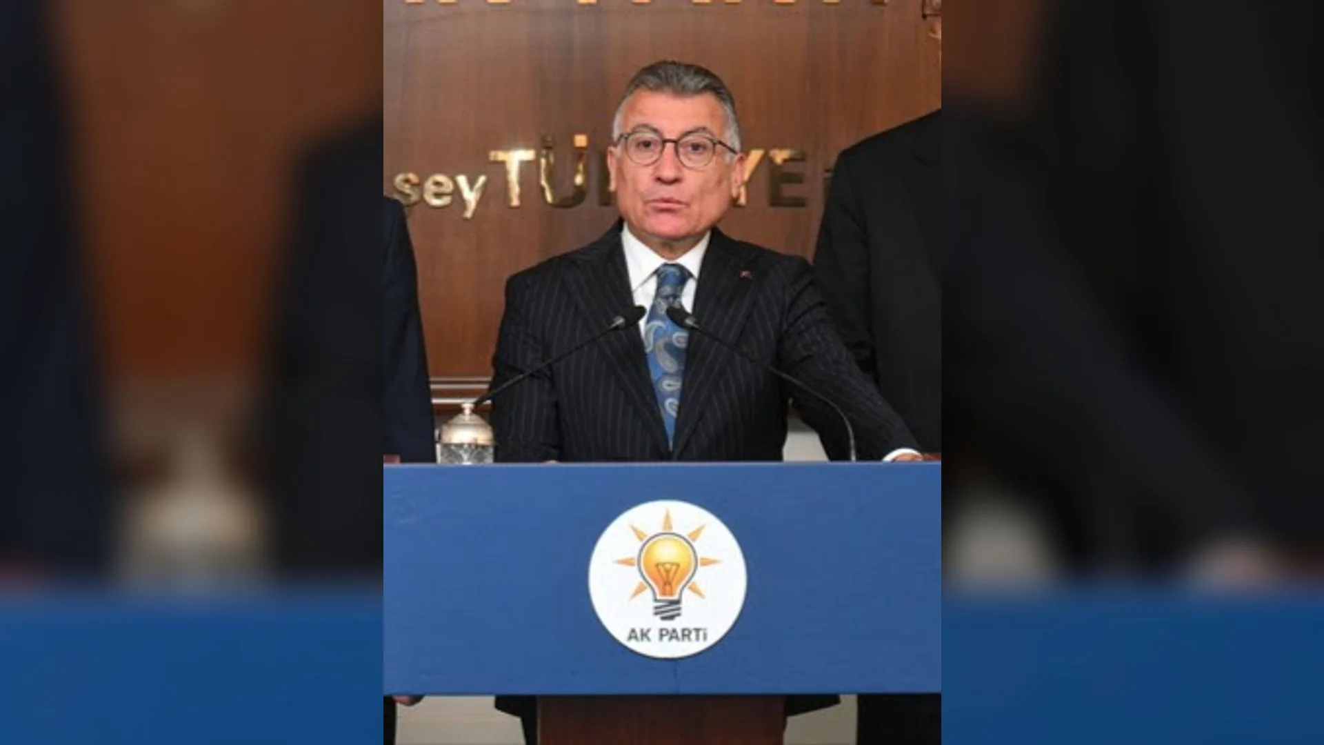 AK Parti logolu mavi kürsüde konuşan gözlüklü, takım elbiseli gri saçlı adam