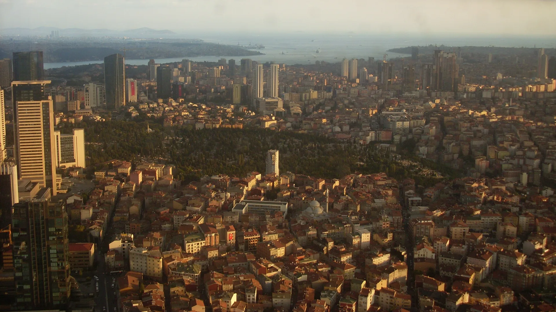 Yüksekten görülen yoğun yapılaşmış bir şehir, gökdelenler, yeşil bir mezarlık alanı ve uzakta boğaz