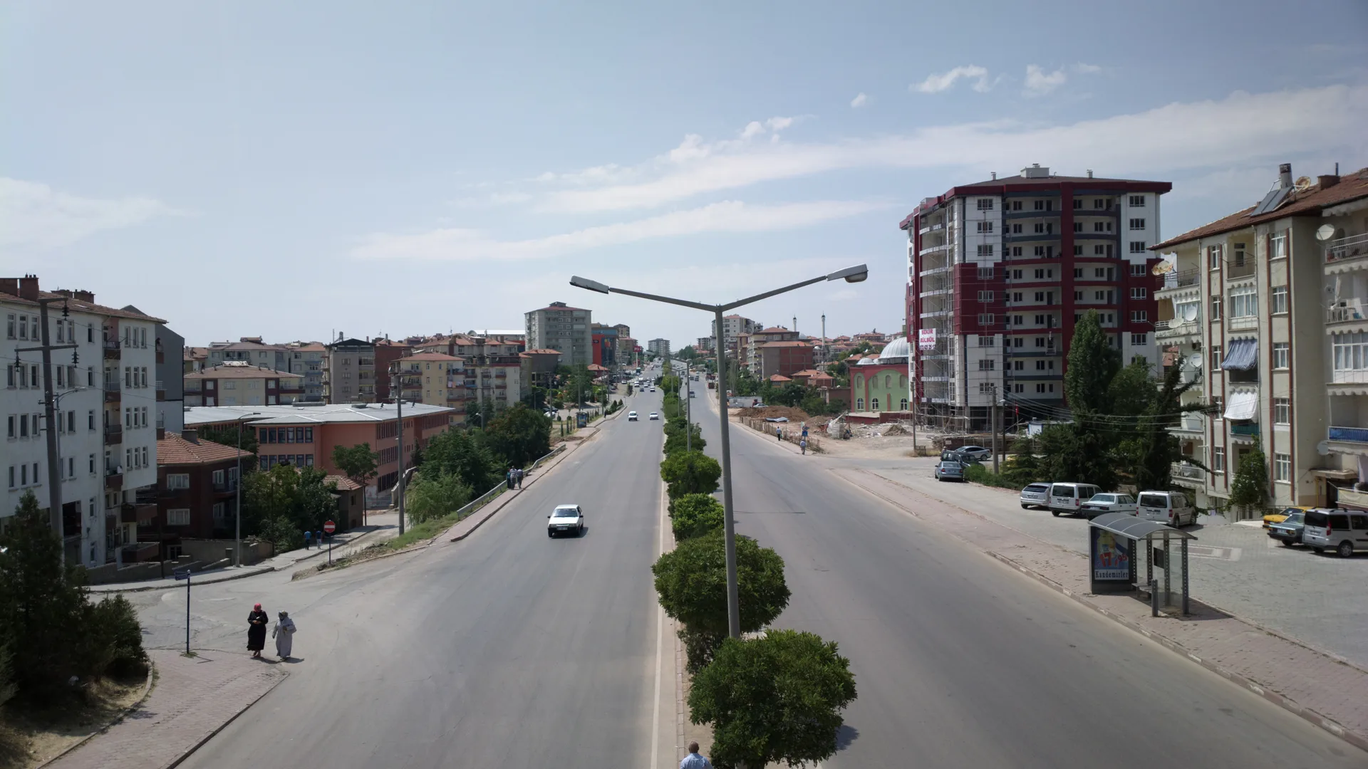Ortası ağaçlı geniş bir cadde, iki yanında apartmanlar ve kırmızı-beyaz yüksek bir bina