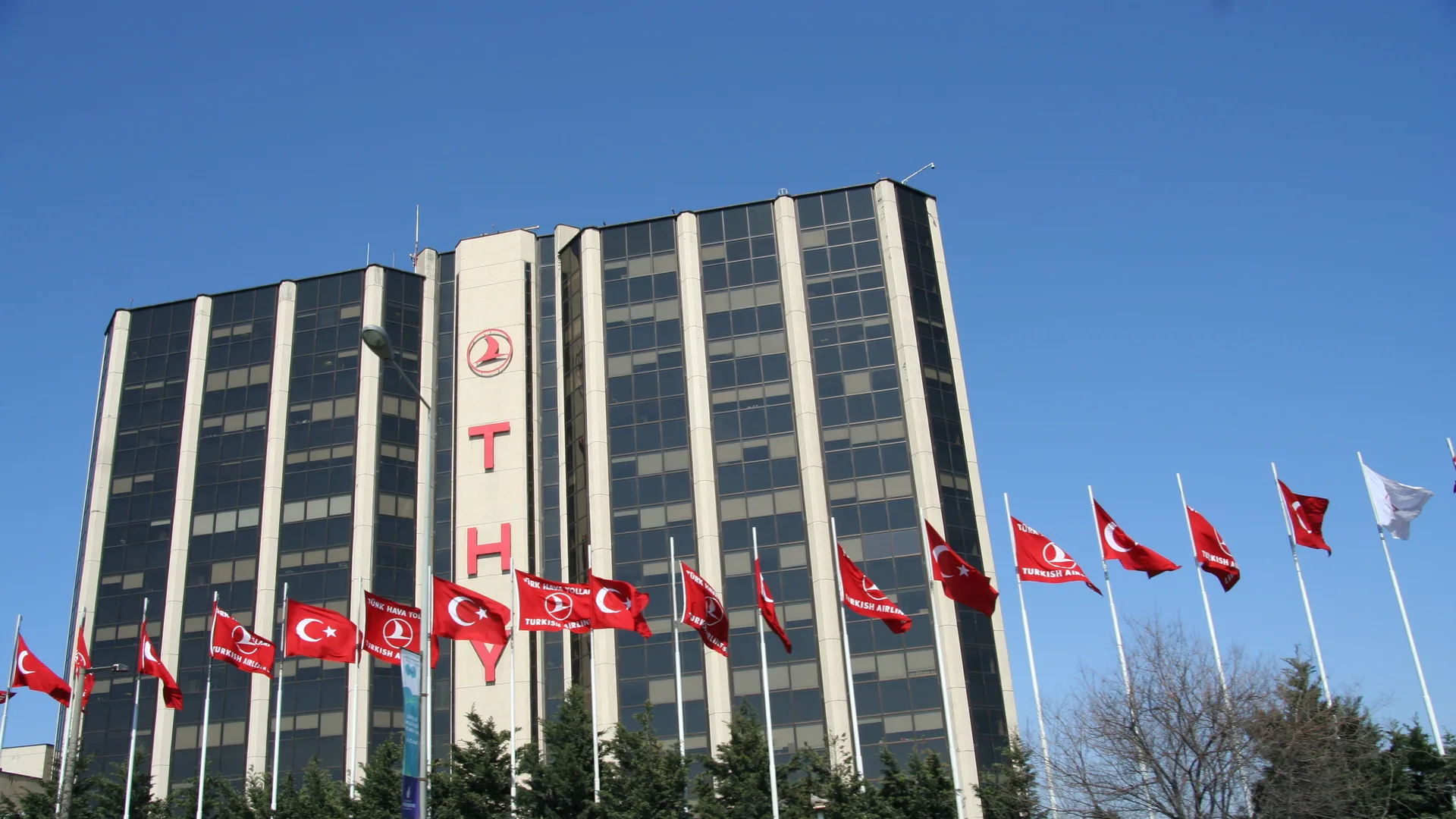 Önünde dalgalanan bayraklar olan, cephesinde THY logosu bulunan cam kaplı yüksek bina