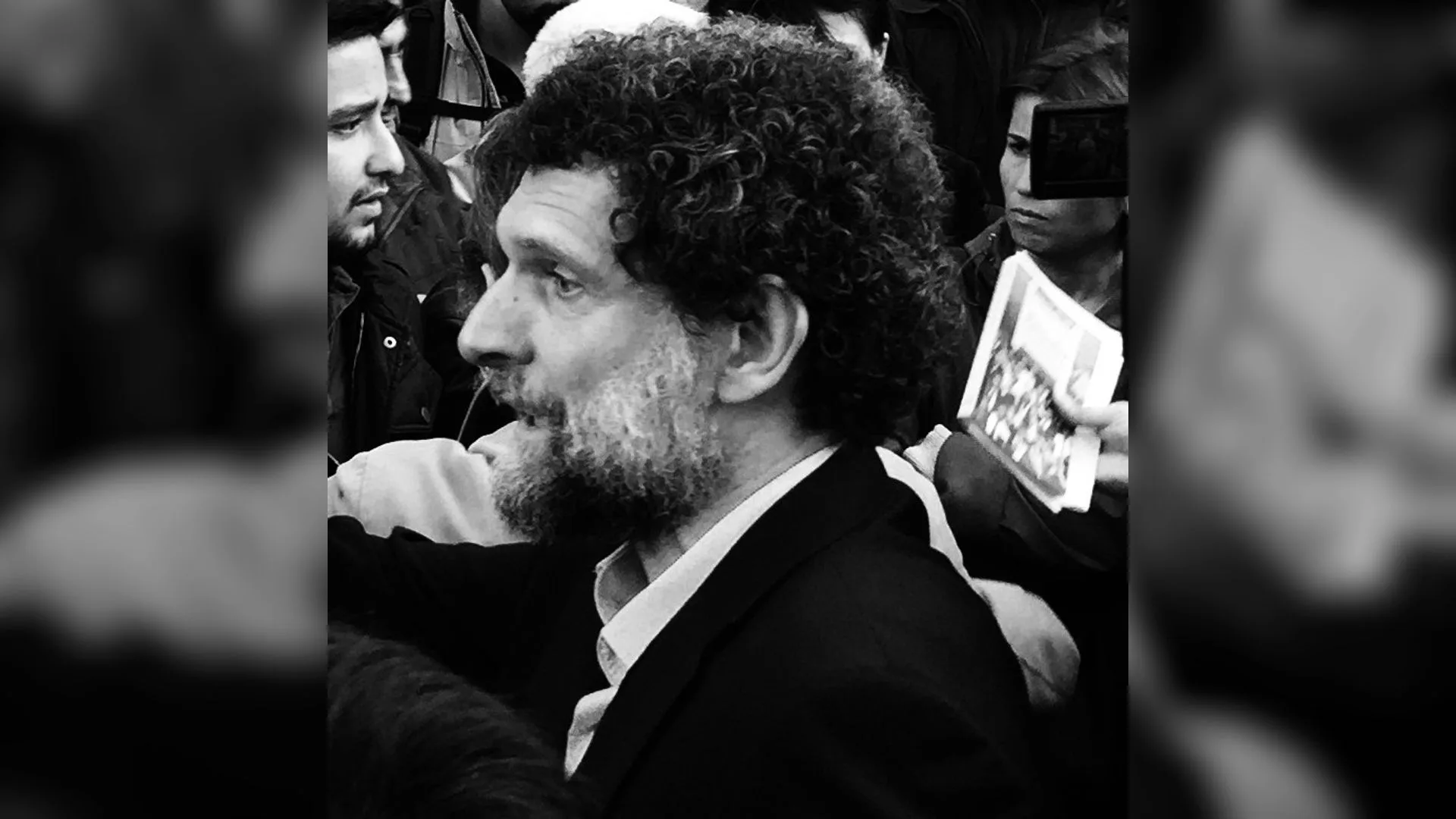 Kıvırcık saçlı, sakallı, ceketli bir adam kalabalığın içinde konuşuyor; siyah beyaz