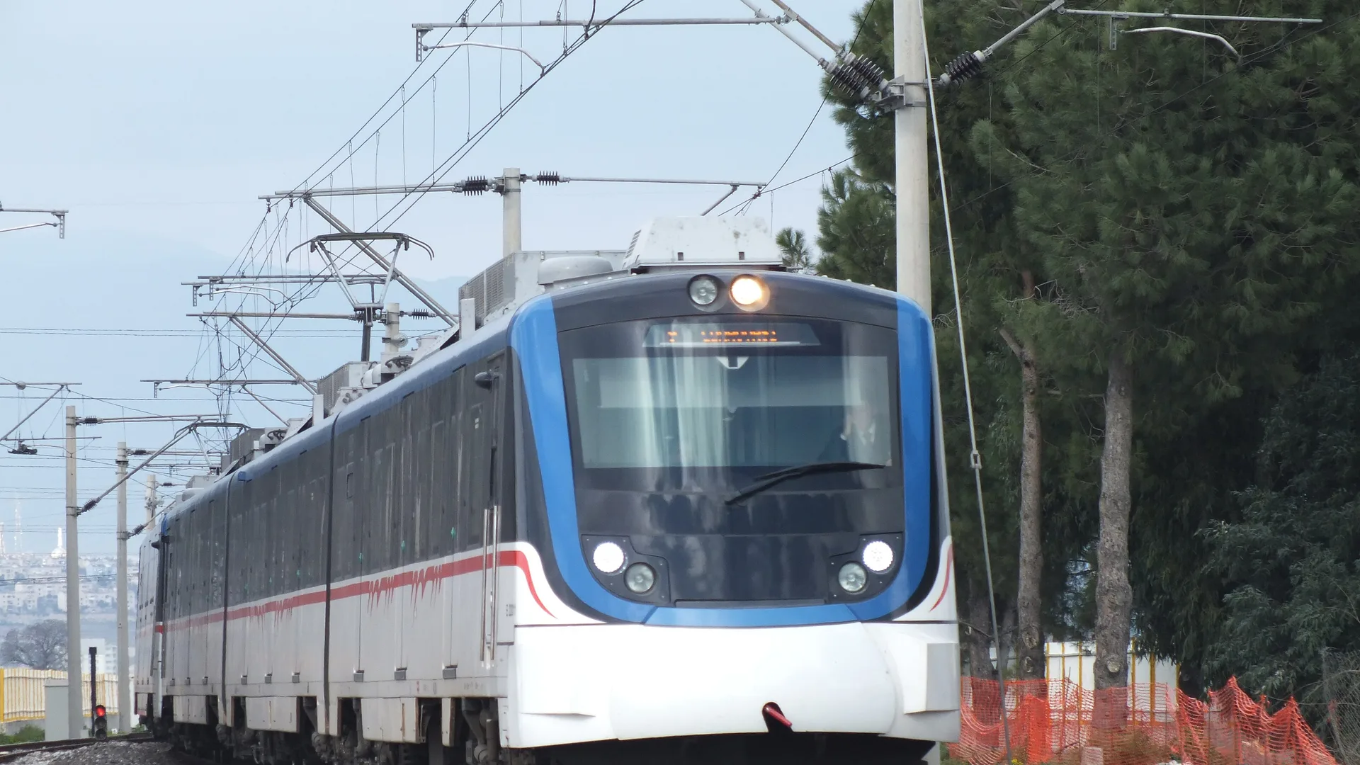 Beyaz-mavi banliyö treni, ağaçlıklı bir hatta raylar üzerinde ilerliyor.
