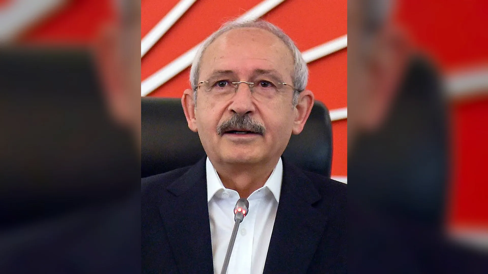 Kırmızı beyaz CHP logolu fonun önünde, mikrofon karşısında konuşan gözlüklü, bıyıklı, beyaz saçlı adam