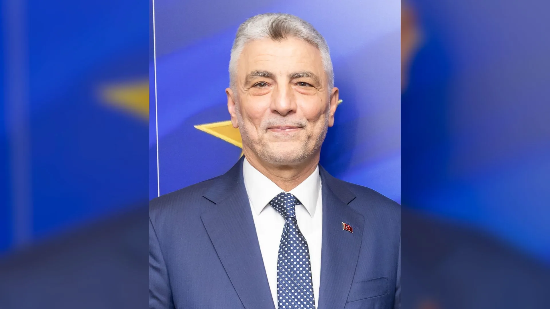Mavi fon önünde lacivert takım elbiseli, puantiyeli kravatlı, gri saçlı gülümseyen bir adam