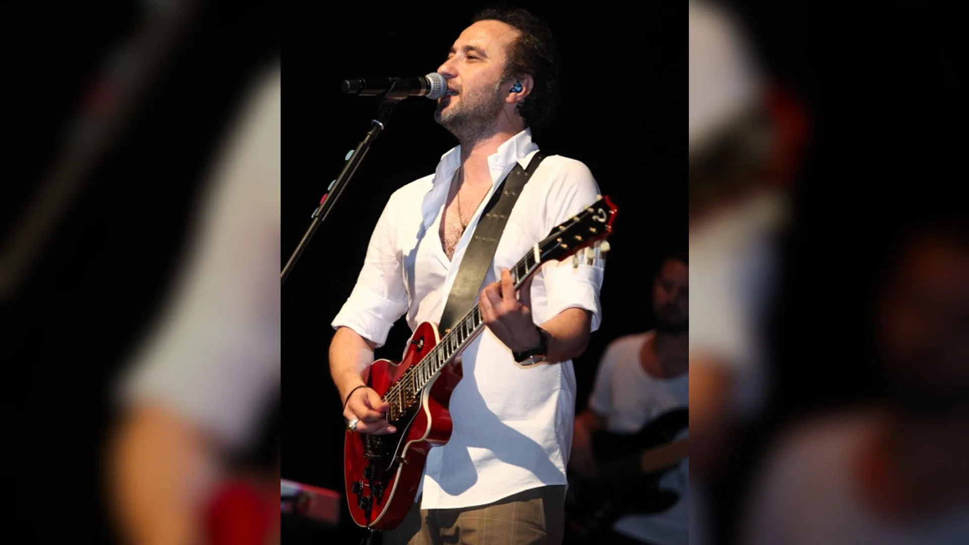Beyaz gömlekli sakallı bir adam sahnede kırmızı elektro gitar çalarak mikrofona şarkı söylüyor