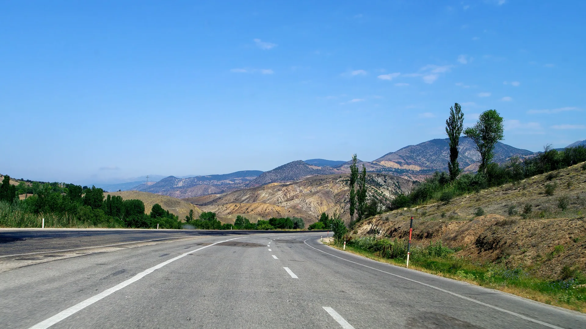 Kurak dağların arasından kıvrılarak geçen boş asfalt yol, kenarında kavak ağaçları