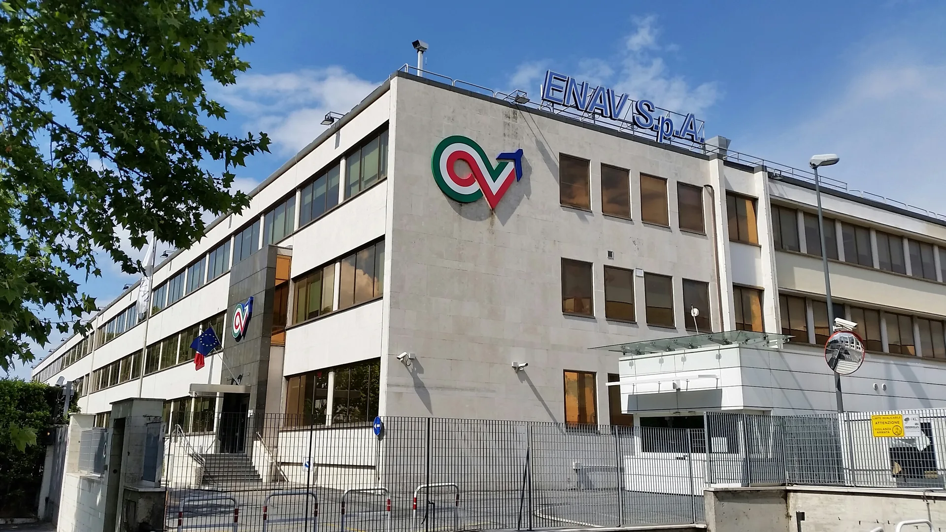 Çatısında ENAV S.p.A. yazısı ve cephesinde renkli logo bulunan, mavi gökyüzü altında çok katlı bir ofis binası