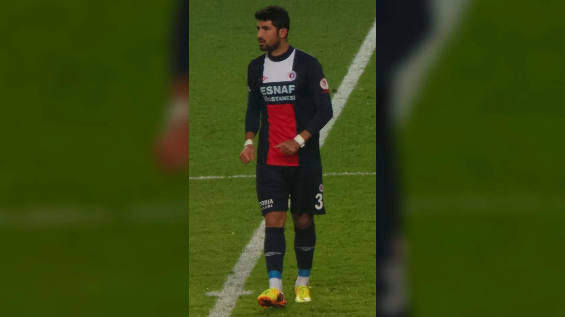 Lacivert-kırmızı forma giymiş, 3 numaralı sakallı bir futbolcu çim sahada duruyor.