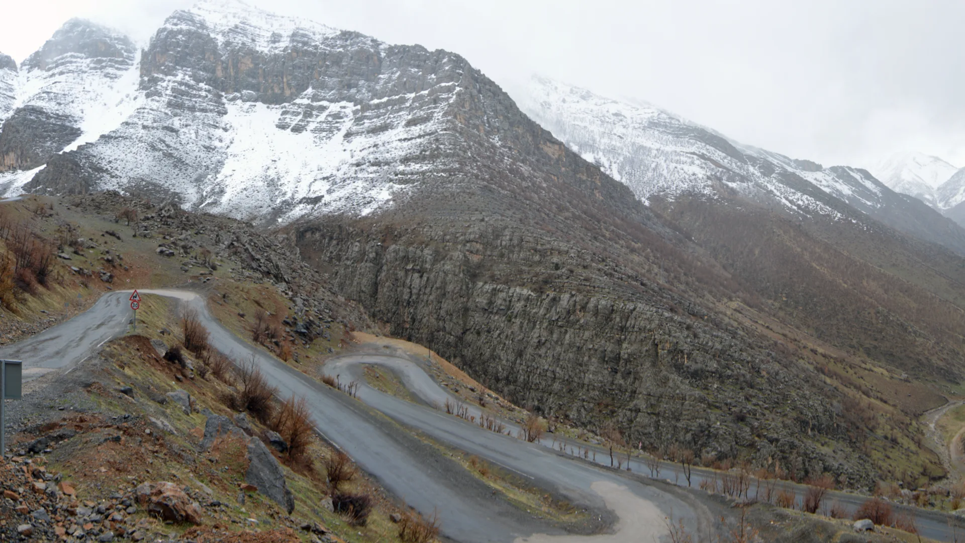 Karlı dağların eteğinde virajlı bir dağ yolu ve yolda duran beyaz bir minibüs