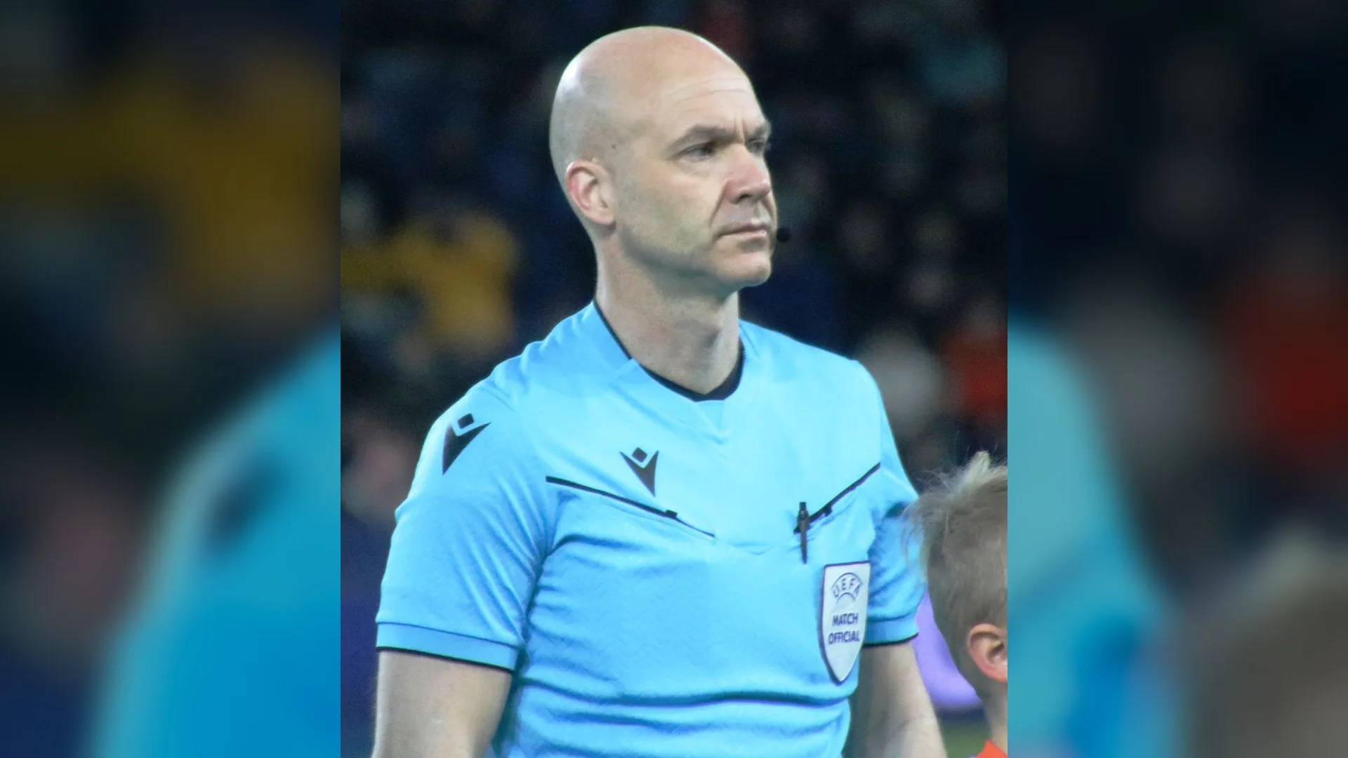 Açık mavi UEFA hakem formalı kel bir adam stadyumda ciddi bir ifadeyle duruyor