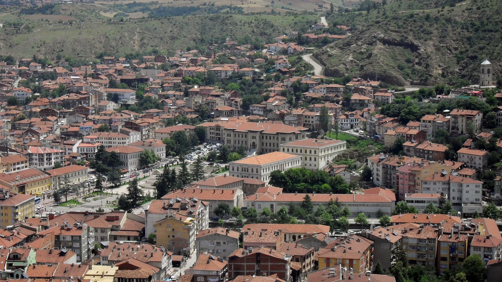 Kırmızı kiremit çatılı evler ve yeşil tepelerle çevrili bir şehir merkezinin yukarıdan görünümü