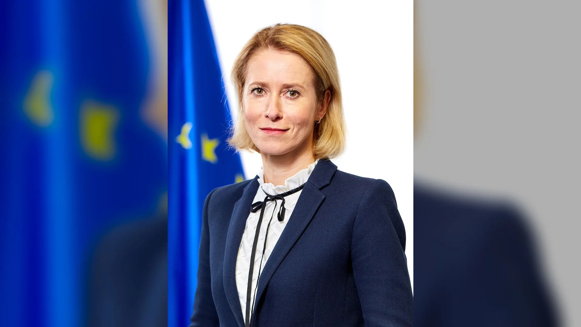 Lacivert ceketli sarışın bir kadın, arkasında Avrupa Birliği bayrağıyla poz veriyor