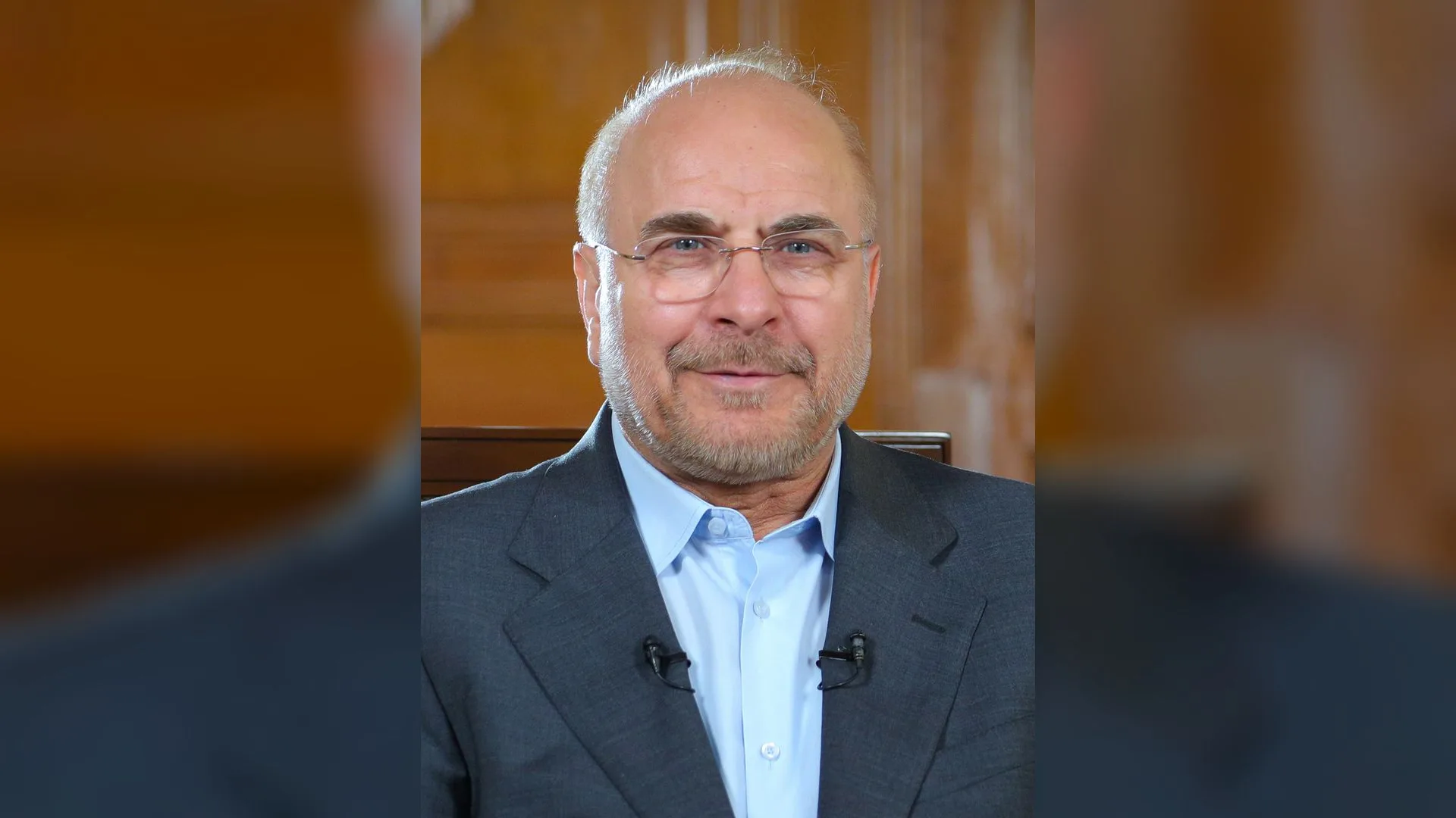 Gözlüklü, kır sakallı, gri ceketli bir adam ahşap duvar önünde gülümsüyor.