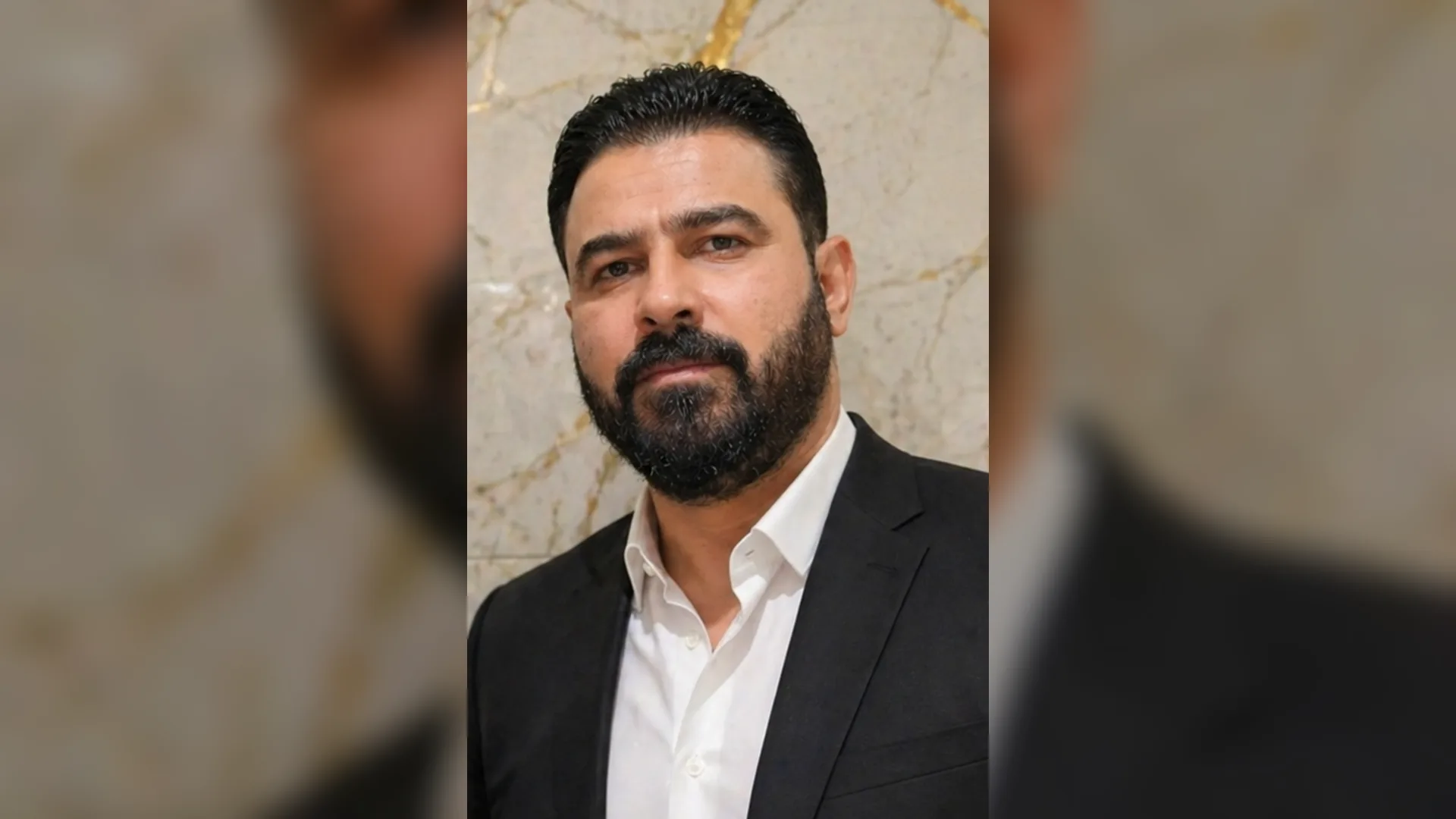 Mermer duvar önünde koyu takım elbiseli, beyaz gömlekli, sakallı bir adamın portresi