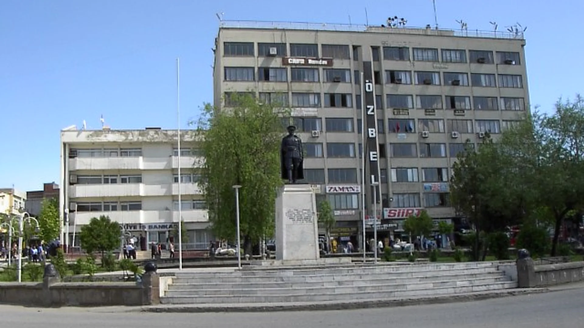 Bina önünde, merdivenli kaide üzerinde asker heykeli; arkada çok katlı binalar ve ağaçlar