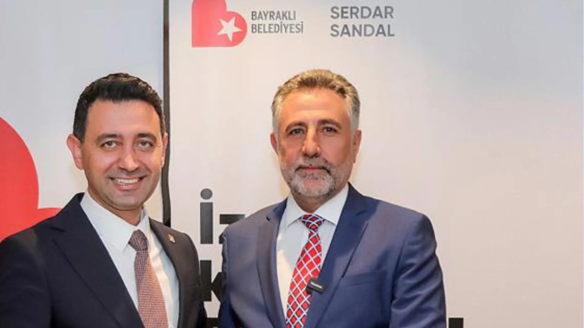 Takım elbiseli iki adam, Bayraklı Belediyesi logolu panonun önünde gülümseyerek poz veriyor.