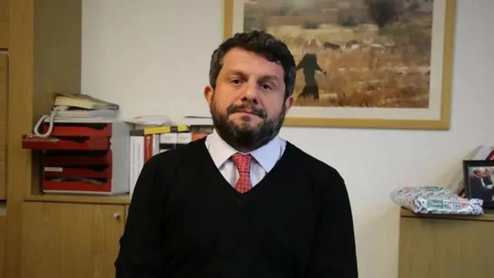 Ofiste oturan, siyah kazaklı, kırmızı kravatlı sakallı adam