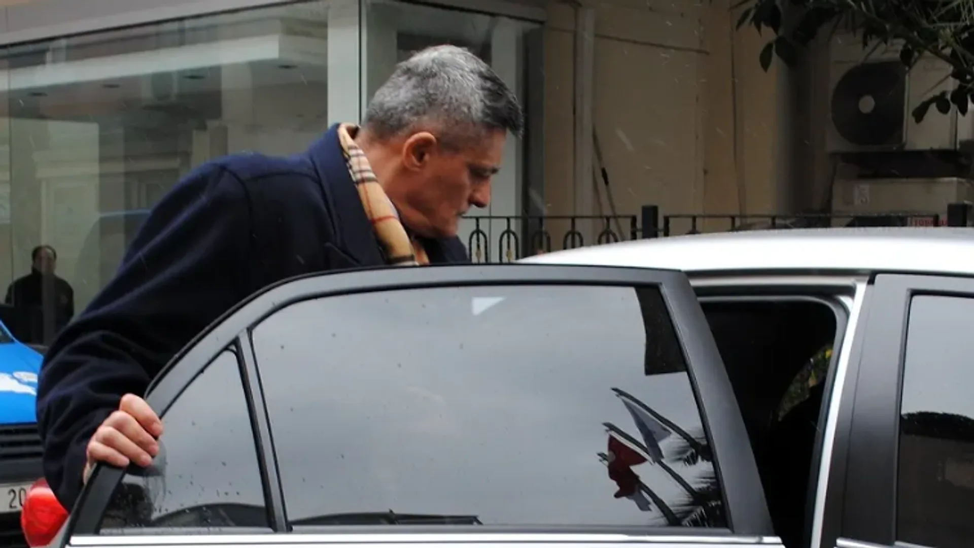 Lacivert paltolu, ekose atkılı, kır saçlı bir adam beyaz bir otomobilin arka kapısından biniyor
