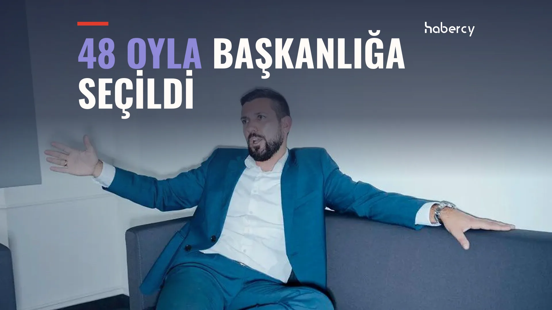 Mavi takım elbiseli sakallı bir adam gri kanepede kollarını açmış oturuyor