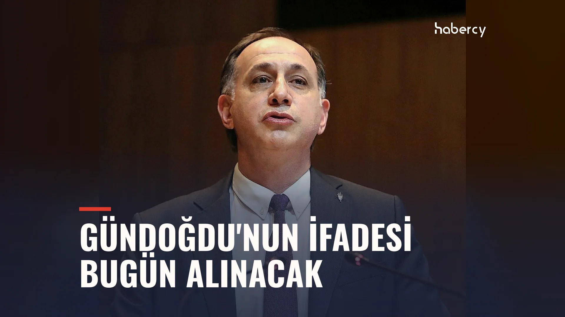 Takım elbiseli, kravatlı bir adam karanlık bir salonda kürsüde konuşuyor
