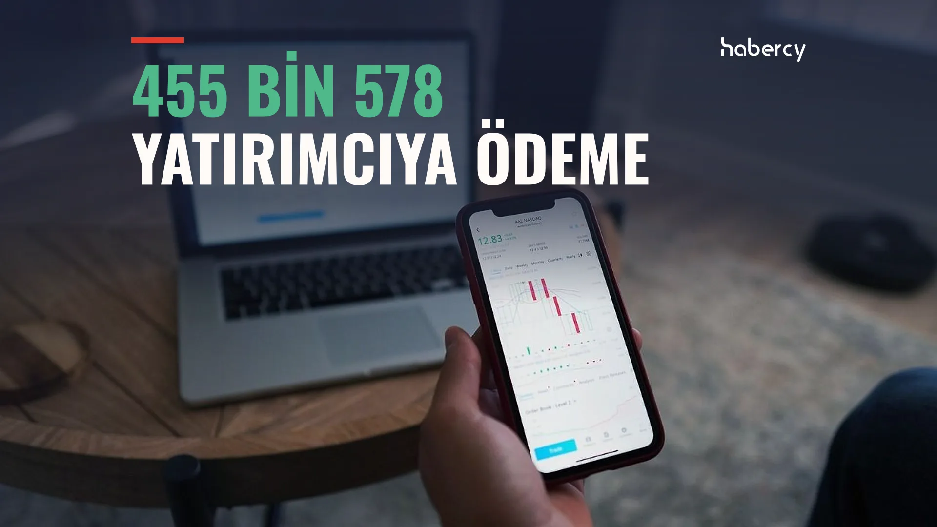 Elinde mum grafiği gösteren telefon tutan biri, arkada masada açık dizüstü bilgisayar
