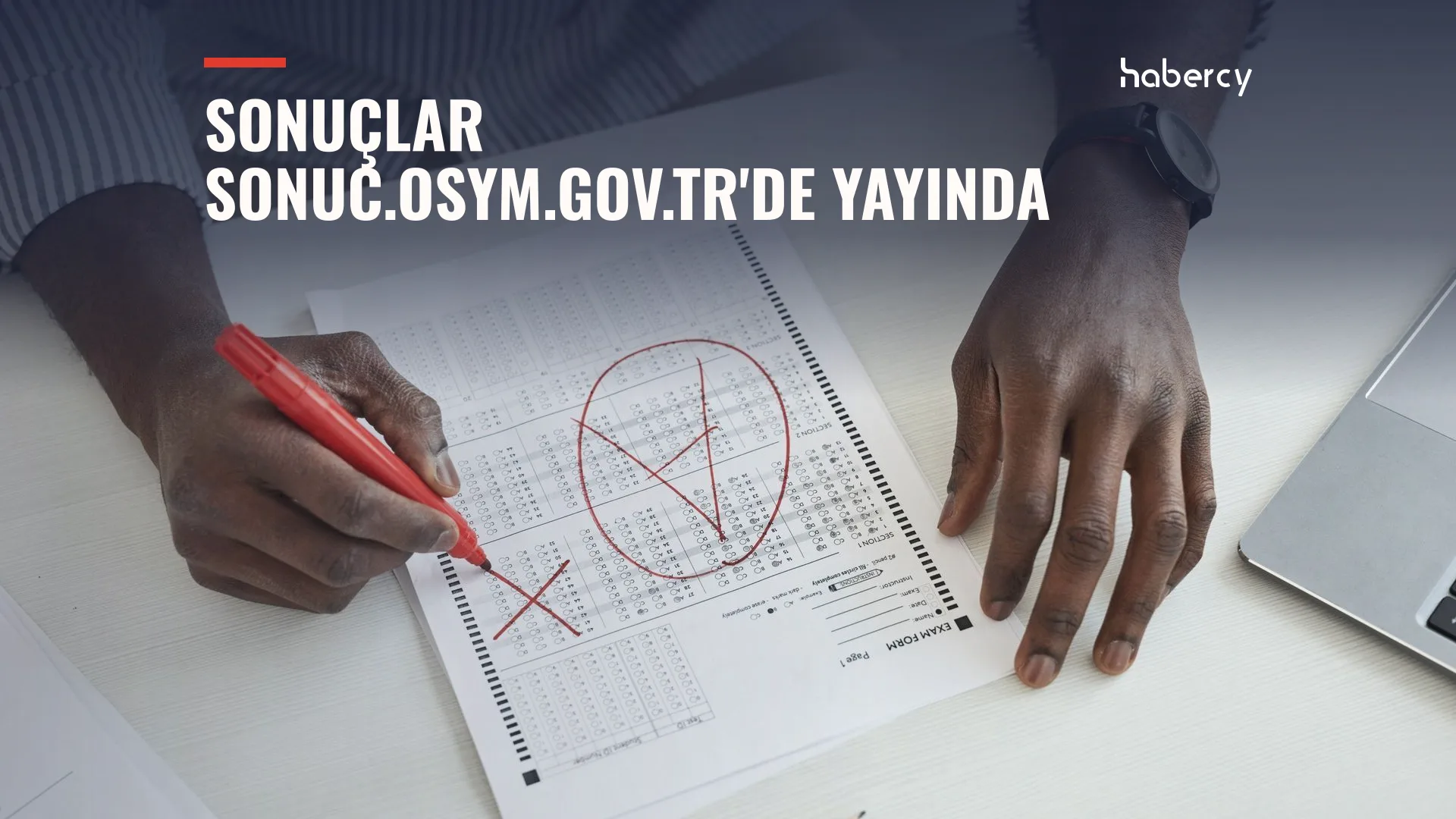 Masada optik cevap kâğıdını kırmızı kalemle işaretleyen eller ve yanda bir dizüstü bilgisayar